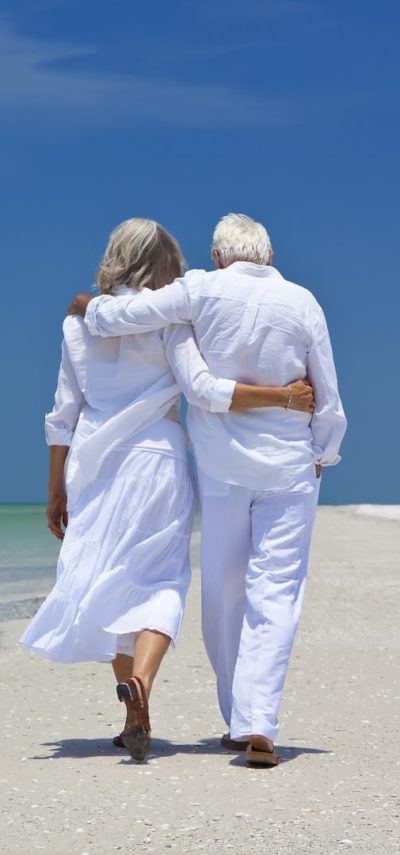 Rear view of a senior man and woman couple walking arms around each other on a deserted tropical beach with bright clear blue sky