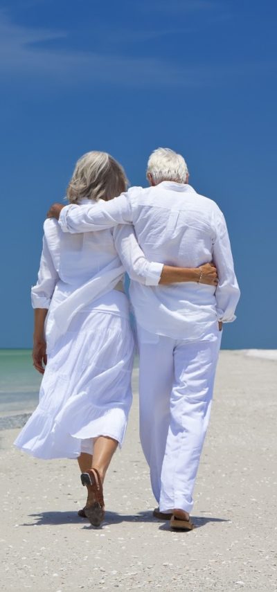 Rear View Of Senior Couple Walking Alone On A Tropical Beach Rear view of a senior man and woman couple walking arms around each other on a deserted tropical beach with bright clear blue sky